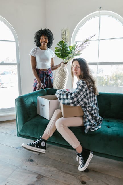 Two women engaged in a home relocation process inside a bright and airy room with large arched windows. One woman, with curly black hair and wearing a white t-shirt and a plaid skirt, is standing behi